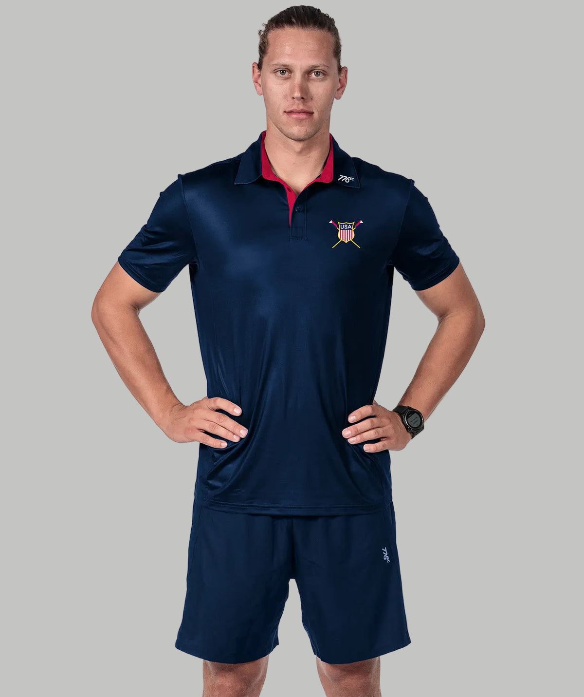 A man modeling a navy blue athletic polo shirt with a red collar and a small crest logo on the chest, paired with matching navy shorts, against a solid gray background.